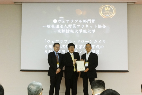 副教授Keiji Emi（中）在2015年10月28日于东京千代田区Sola City会议中心举行的第12届日本电子学习奖上领取可穿戴设备类别奖。
